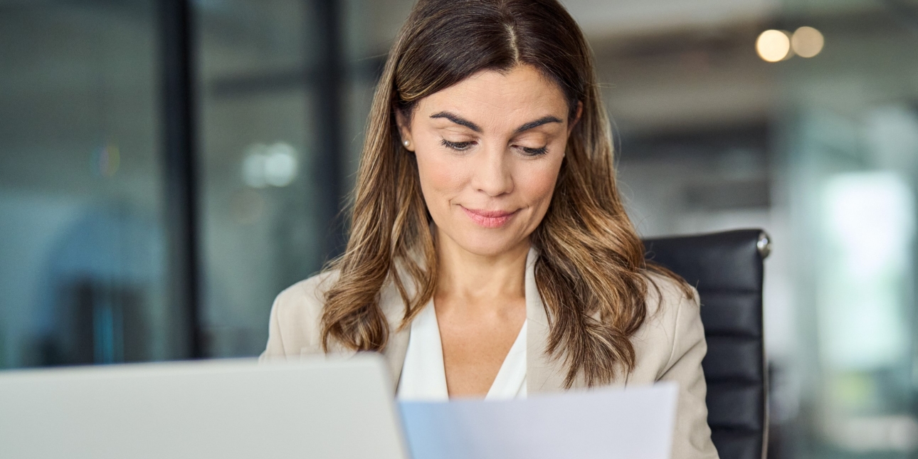 A white woman with mid-length light brown hair wearing a light coloured jacket sits at a laptop and looks at a sheet of paper
